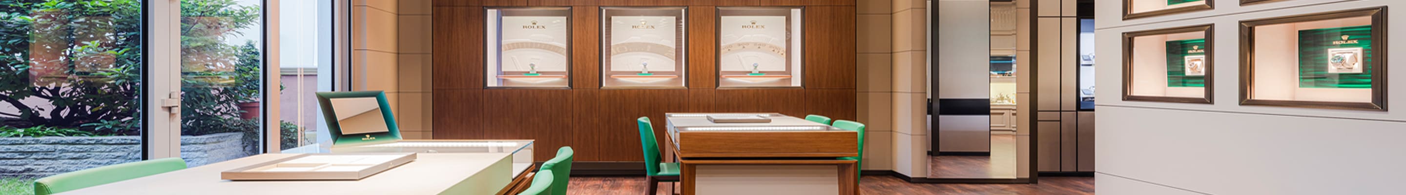Die Rolex Lounge von Juwelier Lorenz ist ein Showroom mit zwei Vitrinen-Tischen und grünen Lederstühlen sowie Schaukästen in der Holzwand.